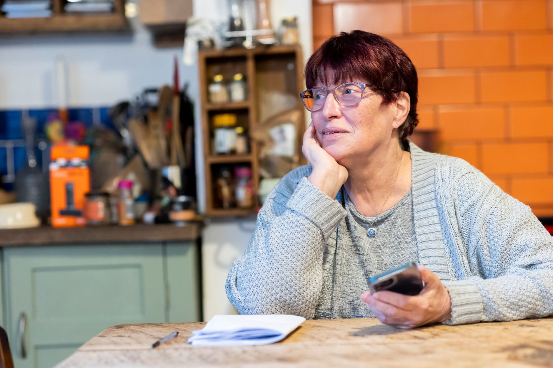 A woman in glasses sitting at a kitchen table with a smartphone and an open notebook