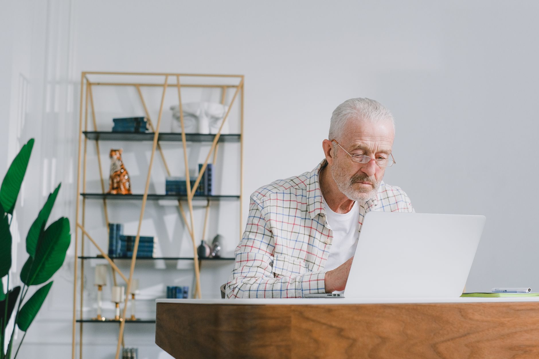 An older man with glasses working on a laptop at a wooden desk in a bright room