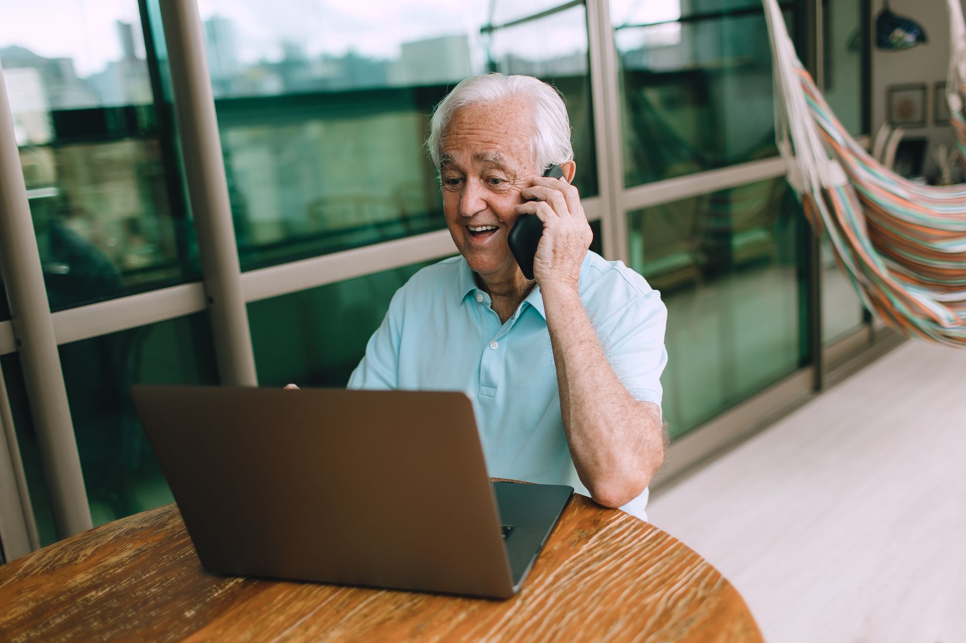 An older man smiles while talking on his phone in front of an open laptop at a wooden table