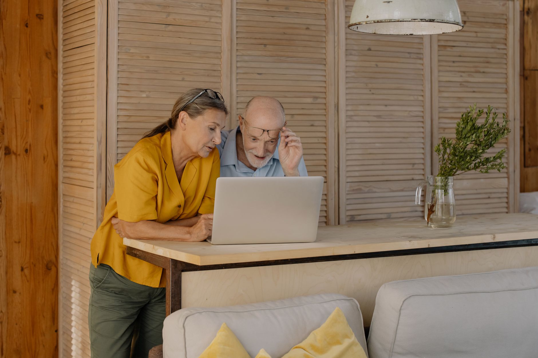 An older couple leaning over a laptop on a wooden counter at home, reading the screen together