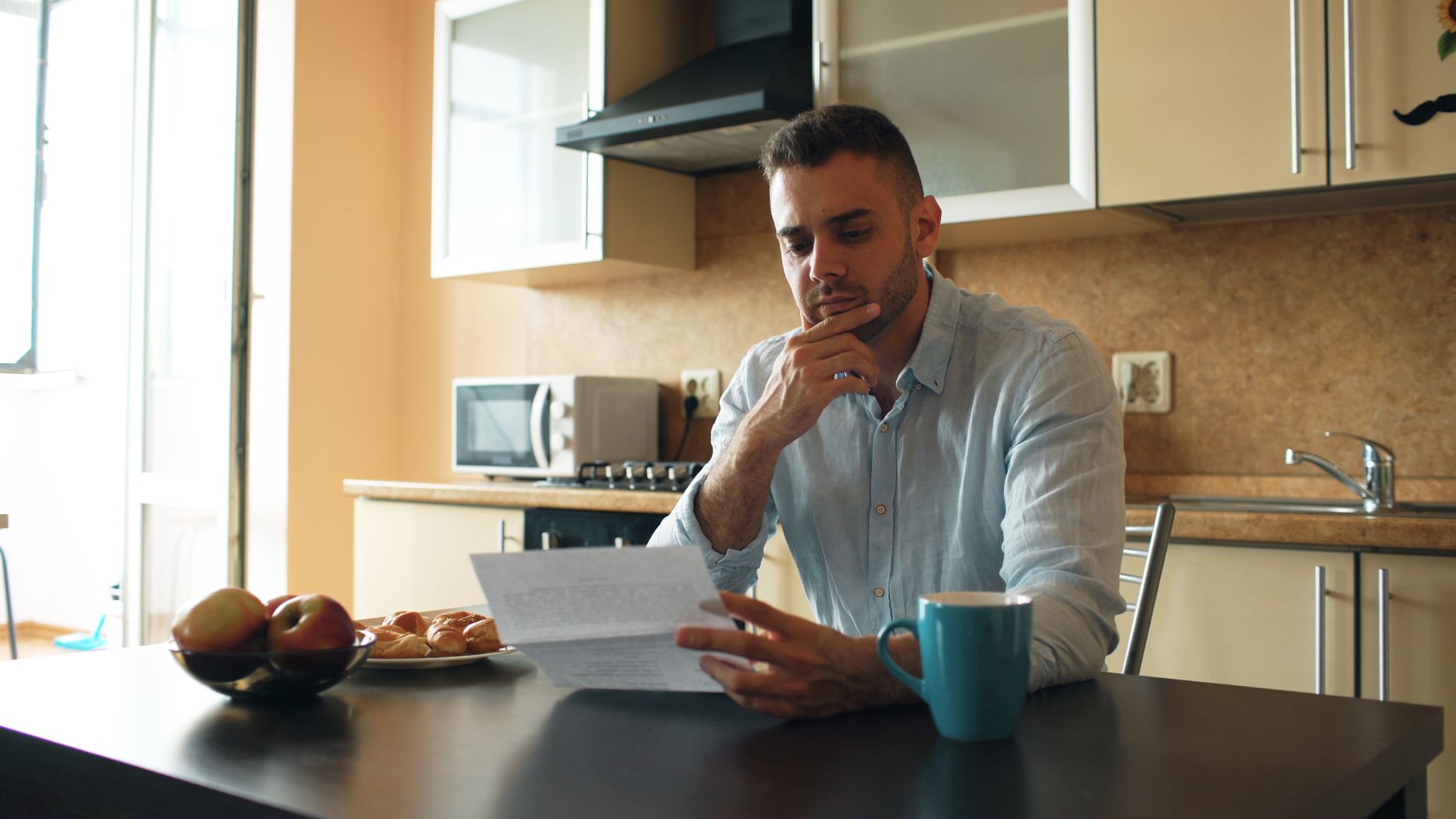 A man sitting at a kitchen table reading a letter with a coffee mug beside him