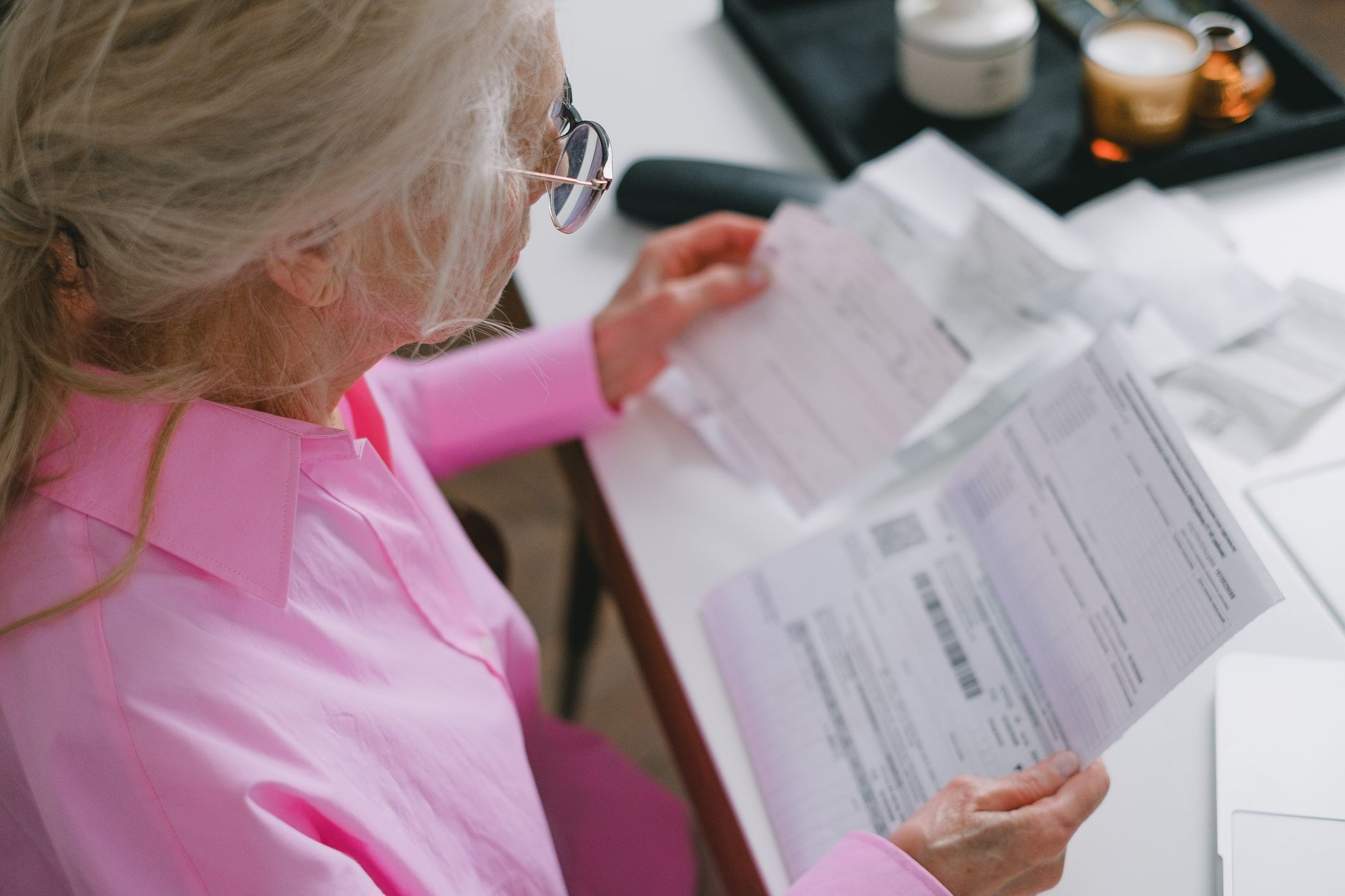 An older woman in a pink shirt reading printed letters and forms at a white table