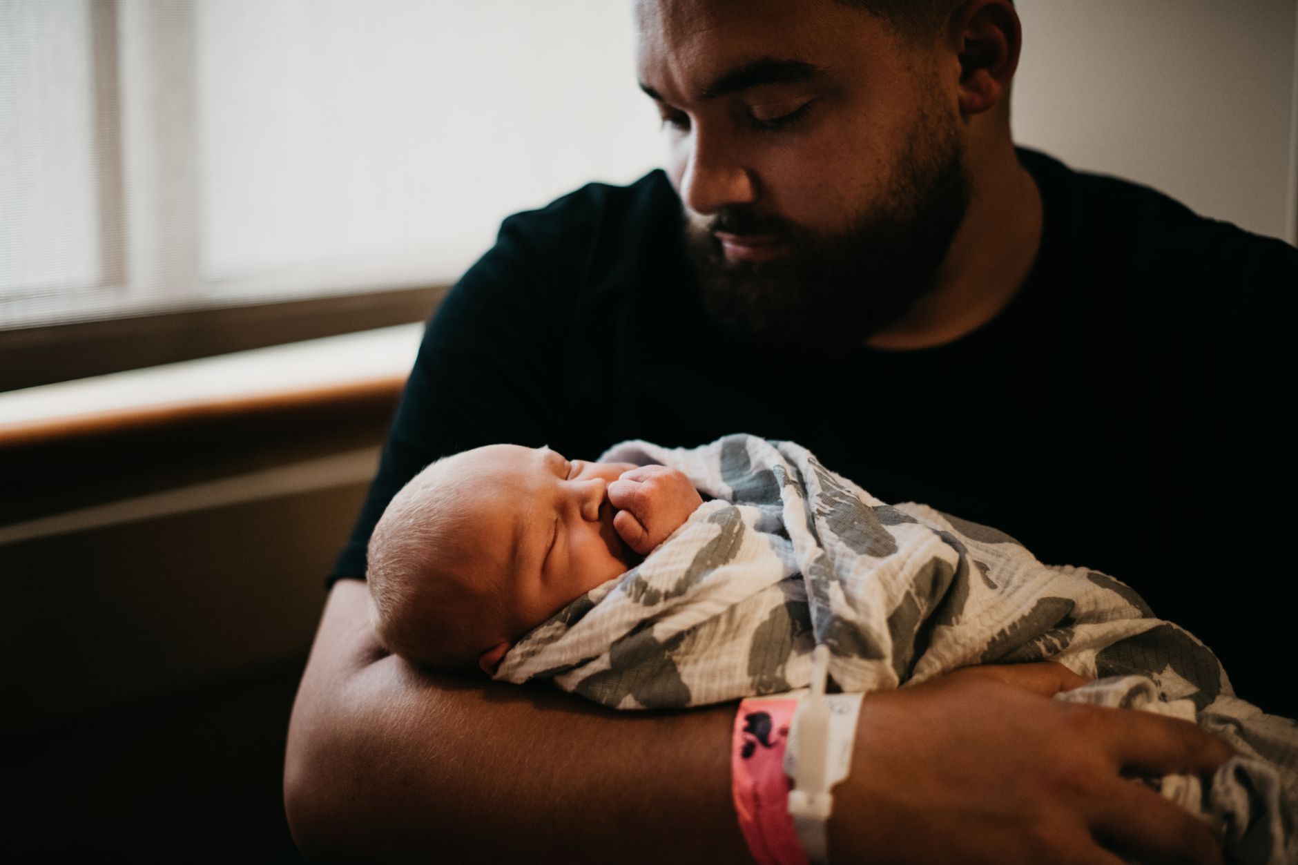 A father in a hospital room cradles his sleeping newborn wrapped in a gray and white blanket