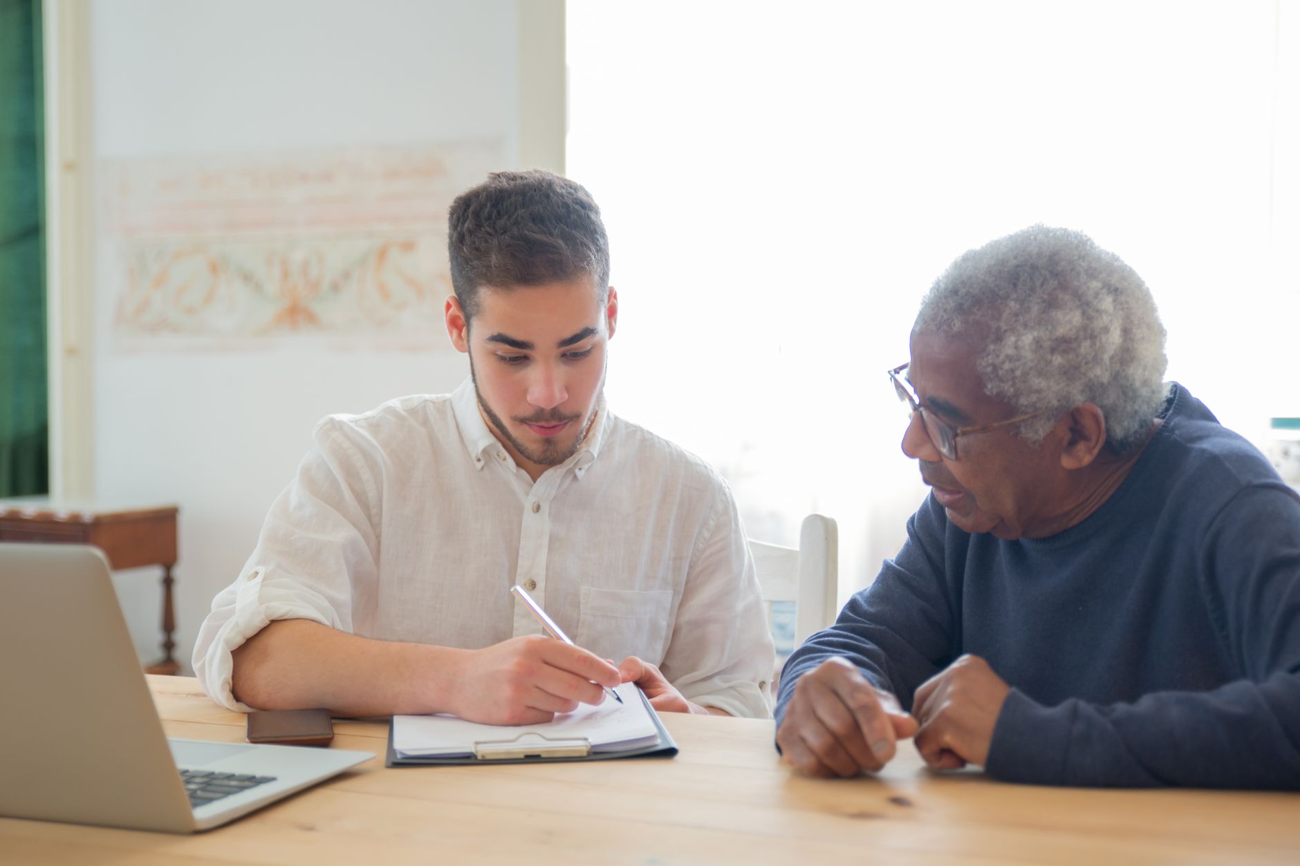 A young man and an older man sitting at a table going over papers on a clipboard next to a laptop
