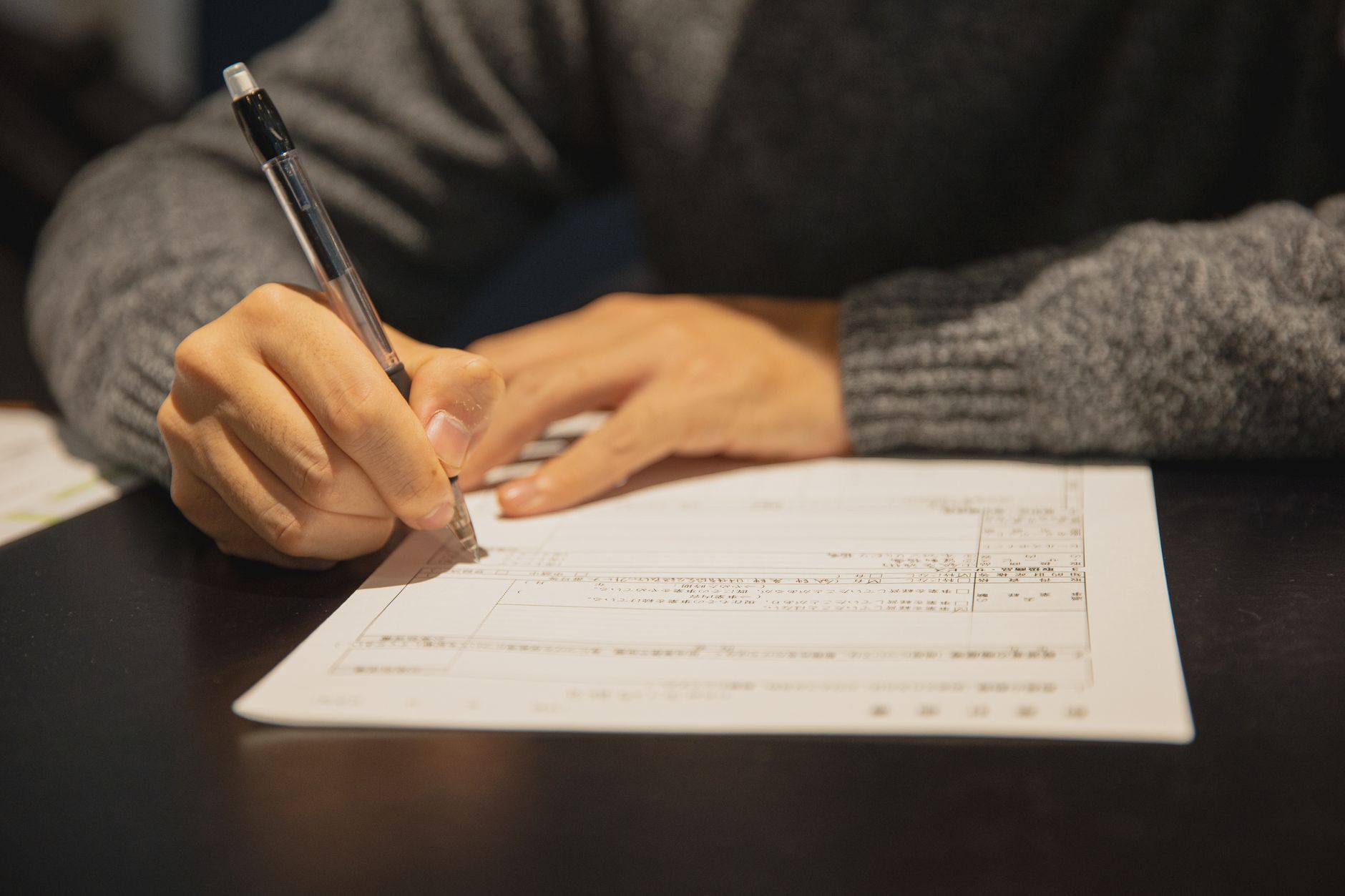 A person in a gray sweater fills out a paper form with a pen at a dark table