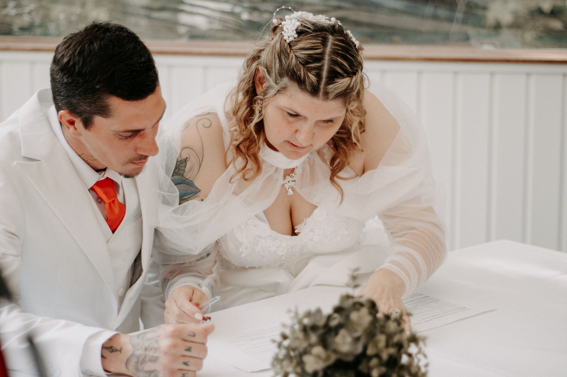 A bride and groom lean over a table together to sign their marriage document
