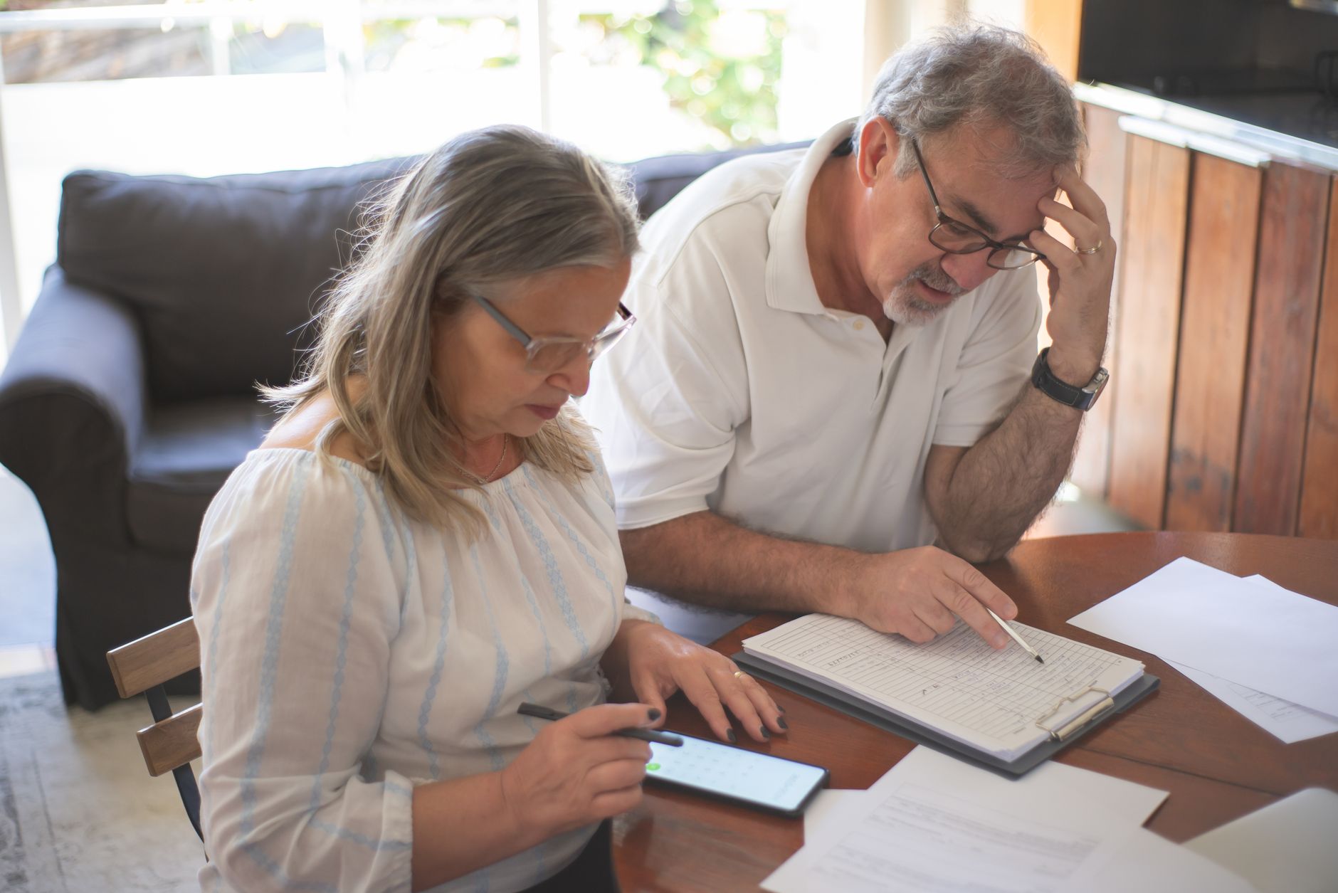 An older couple at a dining table checking a payment log on a clipboard and a calendar on a smartphone