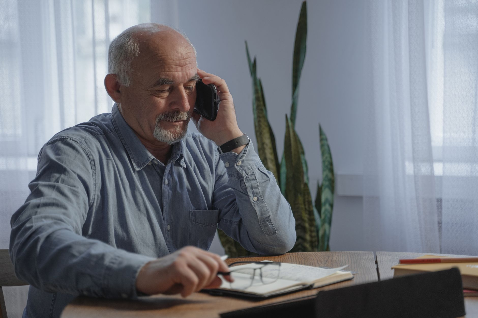 An older man with a gray beard talking on a mobile phone at a wooden table with an open notebook and glasses