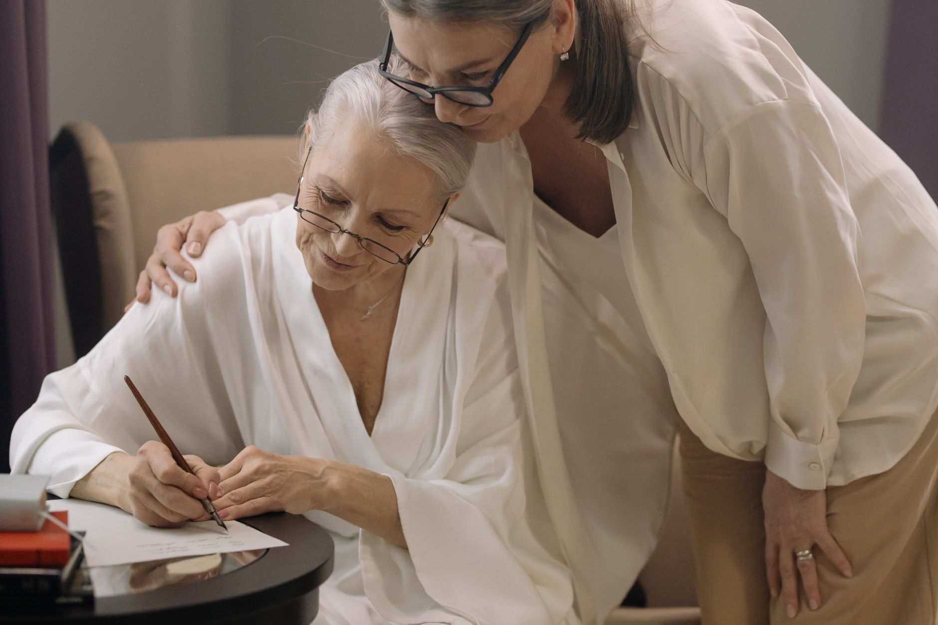 An older woman writing a letter at a small table while a younger relative leans in with a hand on her shoulder