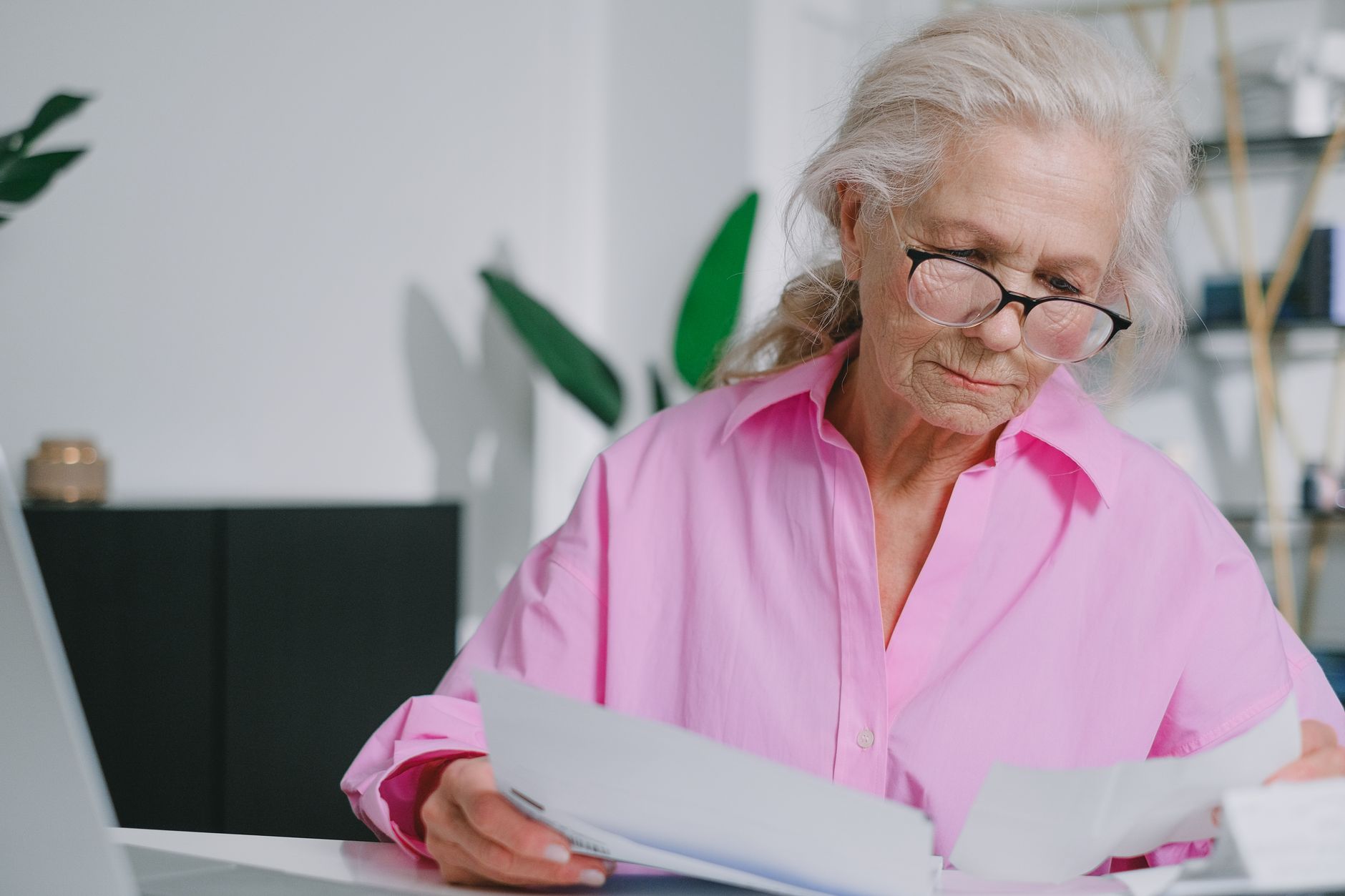 An older woman in glasses reading through printed papers at a white desk